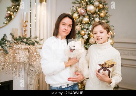 Bella mamma e figlio sorridente che abbraccia un cane bianco felice a casa per le vacanze di natale. Famiglia felice sorridendo e festeggiando il natale con il loro cane divertente. Vacanze di Natale Foto Stock