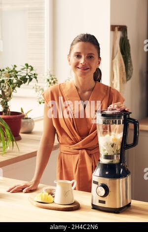 Giovane donna sorridente con frullatore elettrico che ti guarda mentre prepari il frullato per la colazione in cucina Foto Stock
