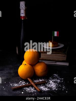 Due bastoncini di cannella sullo zucchero a velo e una piccola piramide di arance con sullo sfondo una bottiglia di champagne e una fetta di torta con la bandiera di esso Foto Stock