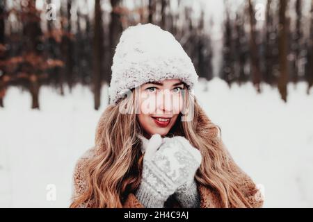 Bella giovane donna in neve fantasia bosco invernale. Ragazza che indossa morbidi guanti, cappuccio e cappotto. Foresta di Natale, alberi su sfondo sfocato. Incrociato Foto Stock