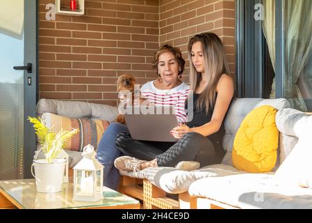 Giovane donna che spiega a una donna anziana con un cucciolo seduto sul divano a casa come utilizzare Internet Foto Stock