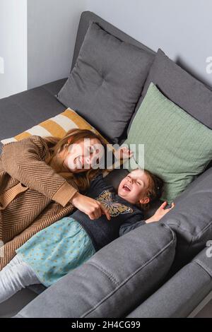 La mamma solletta la figlia mentre si trova sul comodo divano di casa. Risate la famiglia divertendosi insieme Foto Stock