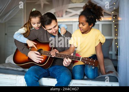 Sorridente padre con bambini multietnici che si divertono e suonano la chitarra a casa Foto Stock