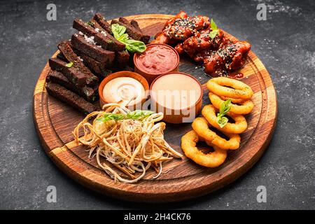 Spuntino con birra su un asse di legno. Anelli di cipolla, crostini, calamari, ali di pollo alla griglia e salse Foto Stock