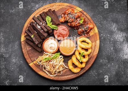 Spuntino con birra su un asse di legno. Anelli di cipolla, crostini, calamari, ali di pollo alla griglia e salse Foto Stock