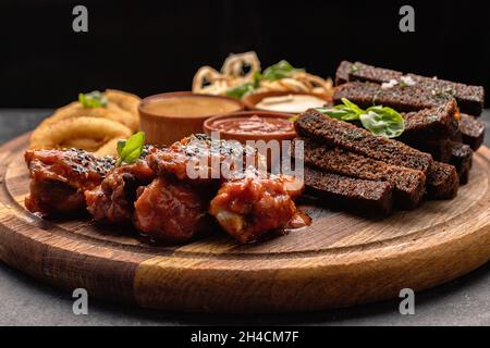 Spuntino con birra su un asse di legno. Anelli di cipolla, crostini, calamari, ali di pollo alla griglia e salse Foto Stock