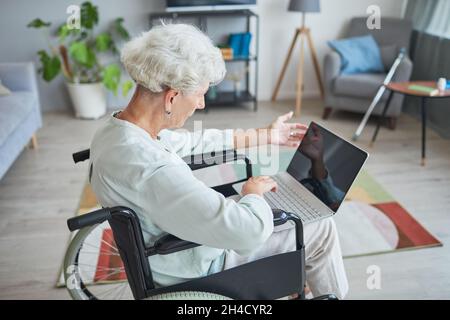 Ritratto ad angolo alto di donna anziana con capelli bianchi in sedia a rotelle usando il laptop a casa Foto Stock