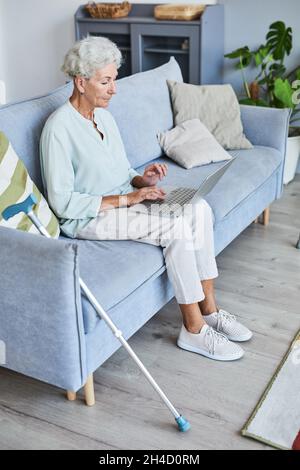 Ritratto verticale a tutta lunghezza di donna anziana con capelli bianchi utilizzando un computer portatile mentre si rilassa sul divano a casa Foto Stock