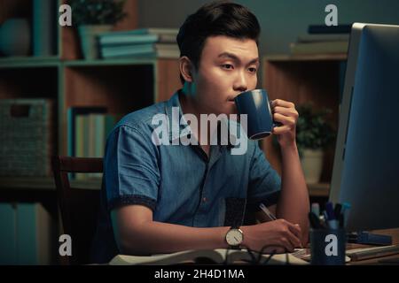 Concentrato giovane specialista asiatico di codifica seduto al tavolo e bere caffè durante il lavoro di notte Foto Stock