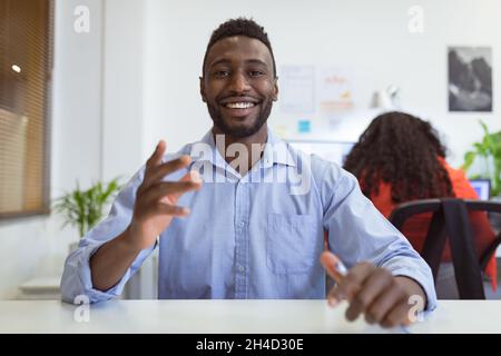 Un uomo d'affari afroamericano sorridente seduto alla scrivania per videochiamare in un ufficio moderno Foto Stock