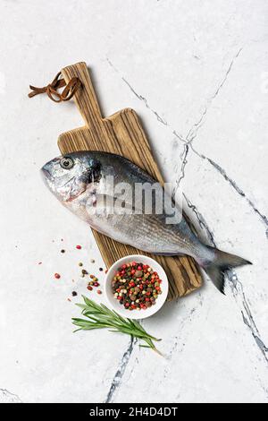 Vista dall'alto di pesce fresco dorado o orata con spezie come pepe, rosmarino su tagliere di legno e sfondo di marmo bianco Foto Stock