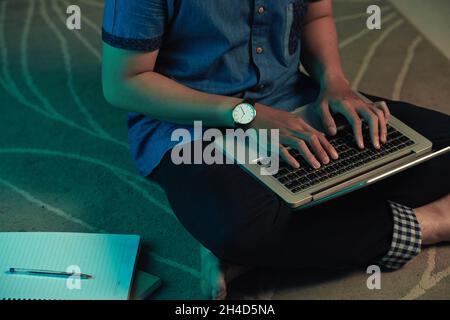 Primo piano di un uomo irriconoscibile in orologio da polso seduto con le gambe incrociate su tappeto e digitando sul computer portatile di notte Foto Stock