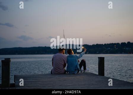 Starnberg, Germania . 23 giugno 2021. Zwei junge Männer sitzen auf dem Großen Steg von Percha und machen ein Selfie mit ihrem handy. Im Hintergrund sieht man das Ufer von Starnberg im Sonnenuntergang. * due giovani uomini si siedono sul grande molo di Percha e prendono un selfie con i loro telefoni cellulari. Sullo sfondo si può vedere la riva di Starnberg al tramonto. (Foto di Alexander Pohl/Sipa USA) Credit: Sipa USA/Alamy Live News Foto Stock