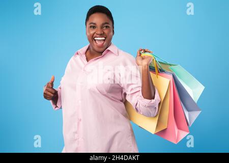 Plus Taglia Black femmina con Shopping Bags gesturing Thumbs-Up, sfondo blu Foto Stock