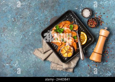 Vista dall'alto dei gamberetti grigliati su padella in ghisa o padella con spezie, limone fritto e aglio su sfondo blu di cemento Foto Stock