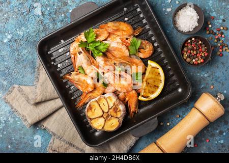 Vista dall'alto dei gamberetti grigliati su padella in ghisa o padella con spezie, limone fritto e aglio su sfondo blu di cemento Foto Stock