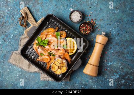 Vista dall'alto dei gamberetti grigliati su padella in ghisa o padella con spezie, limone fritto e aglio su sfondo blu di cemento Foto Stock