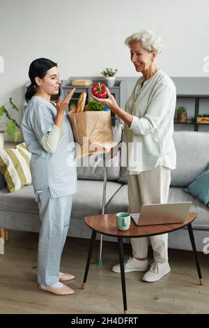 Ritratto verticale a tutta lunghezza di caregiver donna portando generi alimentari a donna anziana Foto Stock
