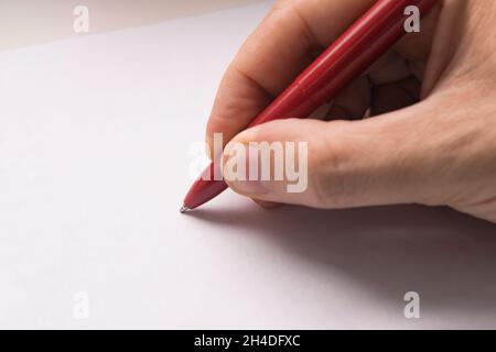 Vista dall'alto della mano maschio con matita su foglio di carta bianco. Su sfondo di cartone. La mano di un uomo scrive con una penna rossa su una superficie di carta bianca. Foto Stock