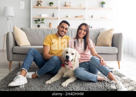 Ritratto a tutta lunghezza di coppia internazionale positiva con il loro cane da compagnia seduto su tappeto morbido a casa Foto Stock