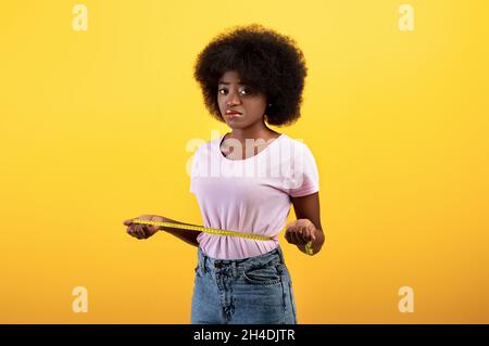 Perdita di peso e concetto di dieta. Triste donna afroamericana che misura la vita con nastro, in piedi su sfondo giallo Foto Stock