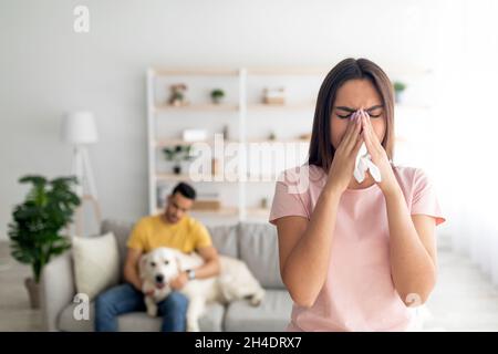 Giovane signora che si sente malbene, avendo freddo, starnutire in carta tissue, il suo ragazzo con cane da compagnia sullo sfondo, copia spazio Foto Stock