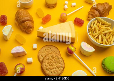 Cibi malsani su sfondo colorato, vista dall'alto. Croissant francese, caramelle su un bastone, caramelle di gelatina, marshmallow, biscotti e macaron su un giallo Foto Stock