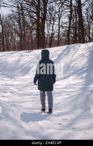 Giovane donna in giacca verde giù è in piedi. Strada invernale in foresta coperta di neve, donna caucasica in abiti caldi passeggiate su bello frosty soleggiato Foto Stock