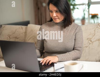 Concetto di ufficio domestico. Donna che lavora su un laptop in remoto sullo sfondo dell'interno del salotto. Donna freelance che lavora da remoto. Lavora da remoto Foto Stock