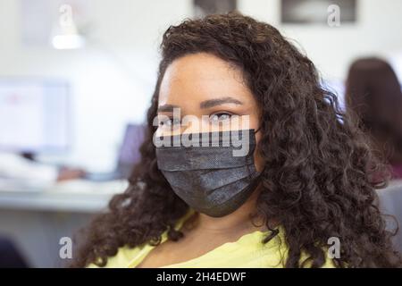 Ritratto di sorridente biraciale businesswoman indossare maschera viso guardando la macchina fotografica in ufficio moderno Foto Stock