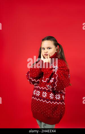 Ritratto di ragazza pensierosa in maglia rossa maglione di Natale con renna che fa il desiderio di Natale, isolato su sfondo rosso Foto Stock