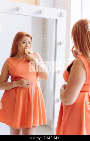 Ansioso donna giovane di grandi dimensioni vestito si leva davanti allo specchio guardando disperatamente al suo riflesso tenendo la mano sullo stomaco e mangiando dolce d Foto Stock