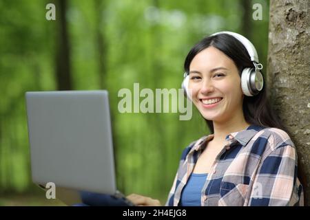 Donna asiatica con computer portatile e cuffie che ti guarda seduto in un parco Foto Stock