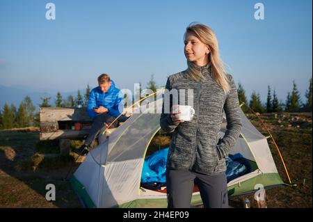 Affascinante escursionista donna che tiene una tazza di caffè e sorride mentre l'uomo è seduto sulla panca e utilizzando lo smartphone. Giovani coppie viaggiatori che riposano all'aperto tenda turistica pulita con cielo blu sullo sfondo. Foto Stock