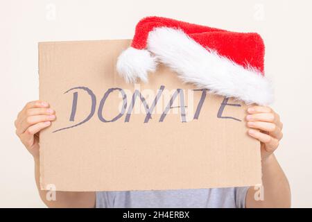 Mani che tengono un pezzo di cartone marrone con iscrizione donate e cappello Babbo Natale. Donazione di Natale, concetto di carità vacanza Foto Stock