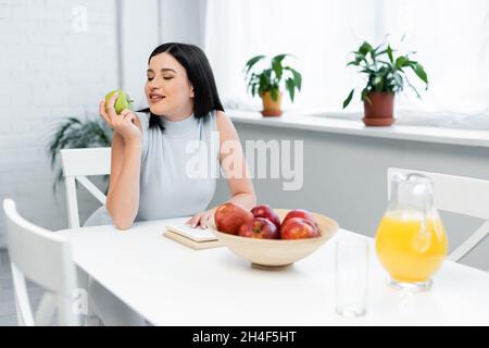 felice donna bruna che tiene mela matura vicino al libro e succo d'arancia sul tavolo da cucina Foto Stock