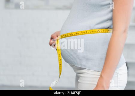 vista parziale della donna incinta che stima le dimensioni della pancia con nastro di misurazione Foto Stock