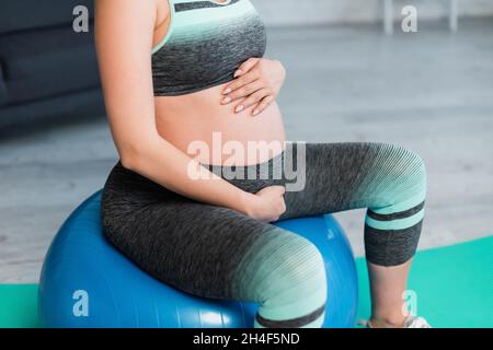 vista parziale della donna incinta che abbraccia il ventre mentre si siede sulla sfera di idoneità Foto Stock