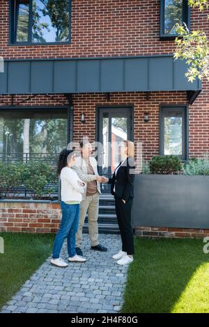 allegro realtor in occhiali scuotendo le mani con uomo sorridente in piedi vicino a moglie afro-americana e casa moderna Foto Stock