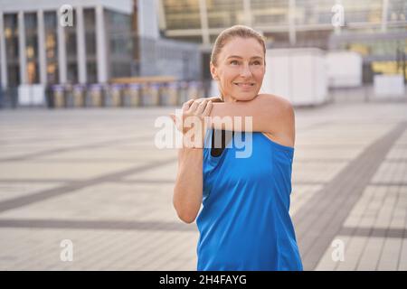 Ritratto di attraente donna di mezza età in abbigliamento sportivo allungando le braccia, in piedi all'aperto, prepararsi per l'allenamento in città Foto Stock