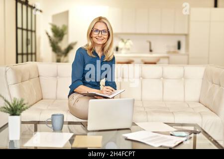 Donna d'affari matura in abbigliamento casual sorridente alla macchina fotografica, lavorando da casa mentre si siede su un divano di fronte al laptop in appartamento moderno Foto Stock