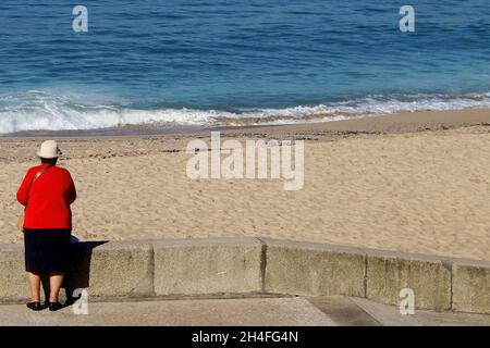 Eine ältere Dame (keine Erkennbarkeit) mit einem roten Blazer, einem schwarzen Rock und weißem Hut steht vor einer Strandmauer, Espinho, Portogallo. Foto Stock