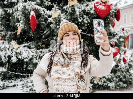 Ritratto di una bella giovane donna felice prendendo un selfie di fronte all'albero di natale in giorno nevoso in città Foto Stock