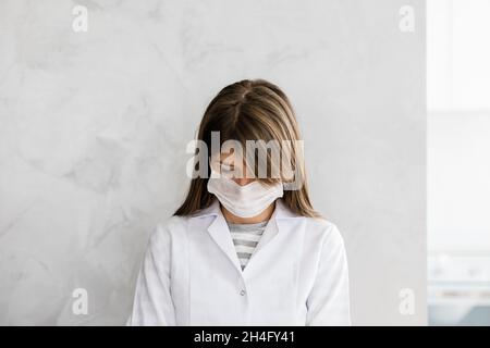 Primo piano di medico caucasico stanco donna con maschera facciale in un abito medico bianco si alza con la testa giù su uno sfondo grigio. Ritratto di femmina doc Foto Stock