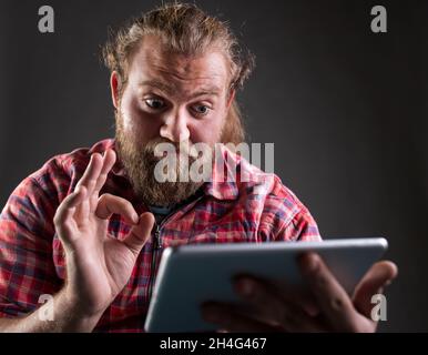 Uomo con barba e capelli lunghi indossare camicia a quadri, tenere un tablet e mostrare il segno ok, gesto soddisfatto, sparare in studio Foto Stock