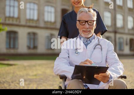 Medico maschile portatore di handicap senior occupato in sedia a rotelle indossando camice da laboratorio e occhiali con giovane infermiere che lo assiste all'aperto Foto Stock