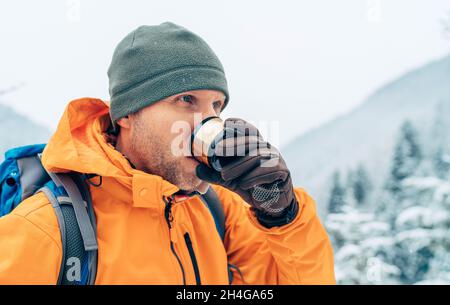 Uomo che beve una bevanda calda da thermos fiasca vestito giacca softshell arancione brillante mentre lui trekking montagna percorso invernale. Persone attive nel natu Foto Stock