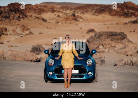 Ragazza in abito giallo godendo di un viaggio su strada in una convertibile attraverso una valle deserta con montagne, Isole Canarie, Tenerife Foto Stock