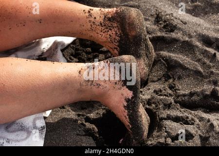 Primo piano delle gambe e dei piedi di una donna nella sabbia, adagiata sulla sabbia nera vulcanica sulla spiaggia.Tenerife. Foto Stock