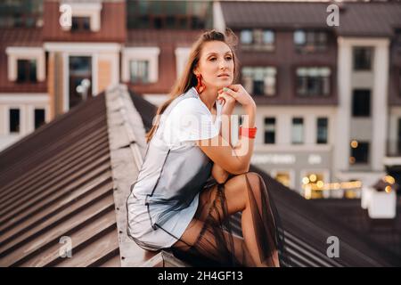 Una ragazza siede sul tetto di una casa in città la sera. Ritratto di un modello in un abito e sneakers Foto Stock